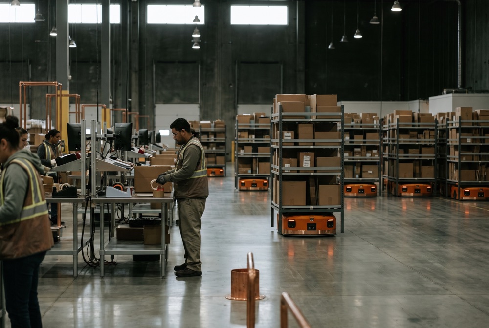 Illustrative warehouse logistics scene with workers, packaged equipment and automated transport robots.