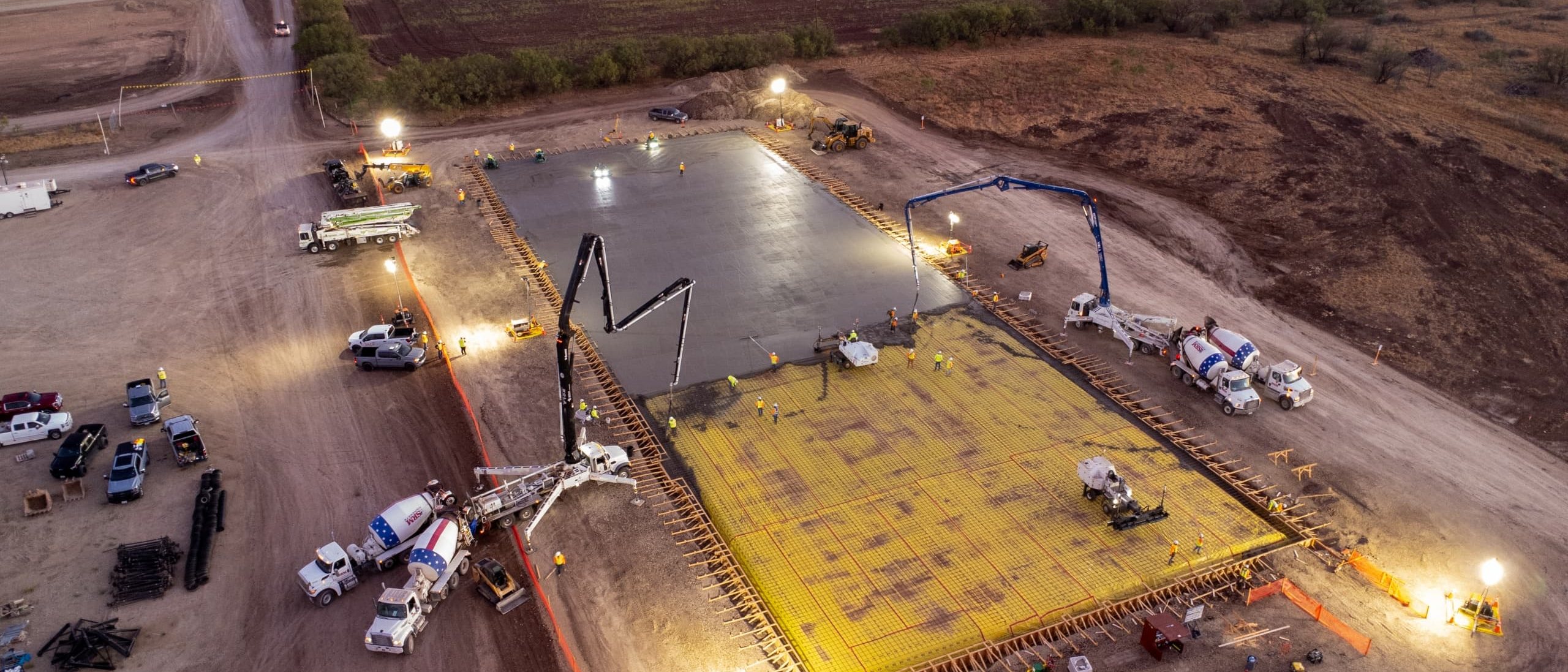 Aerial view at dusk of a DACP Construction crew pouring a data center foundation slab: concrete pumps, mixer trucks and light towers around the rebar mat